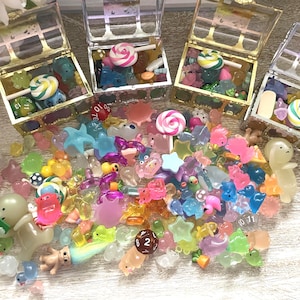 May include: An assortment of colorful miniature charms and candies, including lollipops, dice, and various shaped trinkets. Several clear treasure chests are filled with similar items. The items are in shades of pink, blue, green, and yellow.