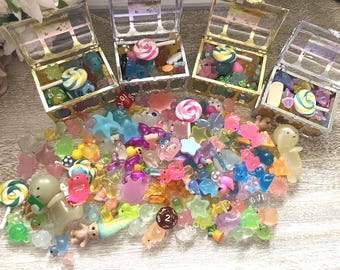 Op de afbeelding: Een assortiment kleurrijke miniatuurbedels en snoepjes, waaronder lolly's, dobbelstenen en diverse gevormde snuisterijen. Verschillende doorzichtige schatkisten zijn gevuld met vergelijkbare items. De items zijn in tinten roze, blauw, groen en geel.