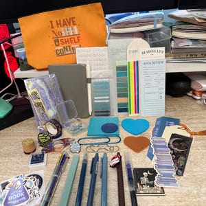May include: An assortment of stationery items, including pens, stickers, notebooks, and bookmarks, are arranged on a light-colored surface. A bright orange pouch with the text "I HAVE NO SHELF CONTROL" is also present. Various colors and designs are visible.