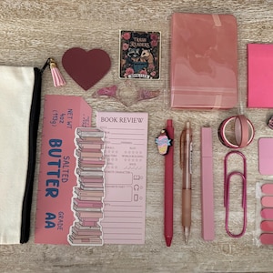 May include: A collection of stationery items including a canvas pouch, a heart-shaped tag, a Trash Readers sticker, a pink notebook, sticky notes, a butter-themed bookmark, a book review notepad, pens, washi tape, a large paperclip, and pins.