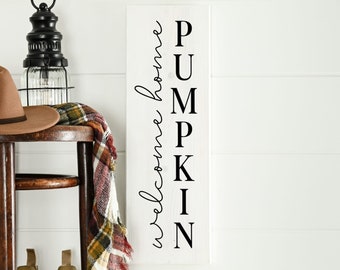 Puede incluir: Un letrero de madera blanco con letras negras que dice "Welcome home PUMPKIN". El letrero está apoyado en una mesa de madera con un sombrero marrón y una bufanda de cuadros en la parte superior.