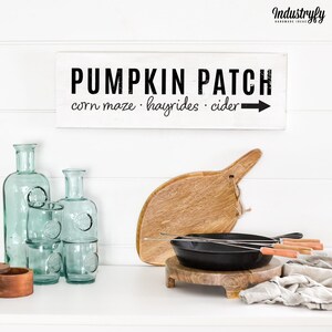 Könnte beinhalten: Ein weißes Holzschild mit schwarzem Text, der "Pumpkin Patch" lautet, mit einem Pfeil, der nach rechts zeigt. Unter dem Text steht "corn maze - hayrides - cider".