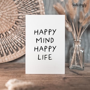 Könnte beinhalten: Eine weiße Karte mit den Worten "HAPPY MIND HAPPY LIFE" in Schwarz, die auf einer Holzoberfläche steht. Die Karte lehnt an einem gewebten Wandbehang und einer Glasvase mit getrockneten Blumen.