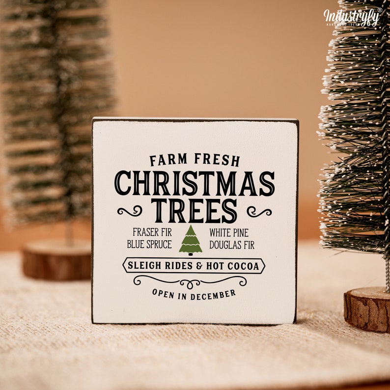 May include: White wooden sign with black text that reads "Farm Fresh Christmas Trees" with a green Christmas tree icon. The sign also says "Fraser Fir, Blue Spruce, White Pine, Douglas Fir, Sleigh Rides & Hot Cocoa, Open in December."