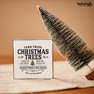 May include: White wooden sign with black text that reads "Farm Fresh Christmas Trees" with a green Christmas tree graphic. The sign also includes the text "Fraser Fir, Blue Spruce, White Pine, Douglas Fir, Sleigh Rides & Hot Cocoa, Open in December".