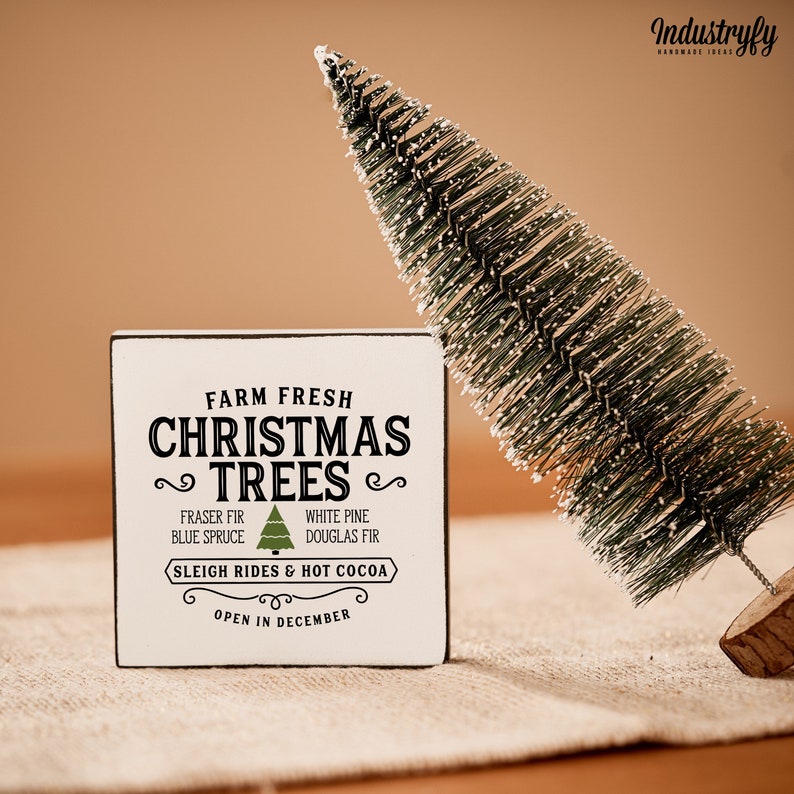 May include: White wooden sign with black text that reads "Farm Fresh Christmas Trees" with a green Christmas tree graphic. The sign also includes the text "Fraser Fir, Blue Spruce, White Pine, Douglas Fir, Sleigh Rides & Hot Cocoa, Open in December".