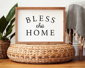 May include: A framed wooden sign with the words "BLESS this HOME" in black lettering. The sign is set on a woven, circular ottoman. A potted plant and a patterned throw blanket are visible in the background, creating a cozy home decor scene.