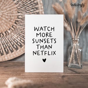 Könnte beinhalten: Eine weiße Karte mit dem Text "WATCH MORE SUNSETS THAN NETFLIX" in Schwarz, mit einem kleinen Herz darunter. Die Karte liegt auf einer Holzoberfläche, mit einem geflochtenen Korb und einer Vase mit Trockenblumen im Hintergrund.