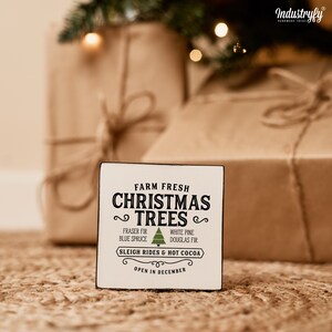 May include: A white wooden sign with black text that reads "Farm Fresh Christmas Trees" with a green Christmas tree icon. The sign also includes the text "Fraser Fir, Blue Spruce, White Pine, Douglas Fir, Sleigh Rides & Hot Cocoa, Open in December."