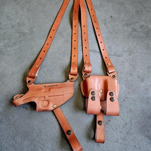 May include: A brown leather shoulder holster with two pouches and a gun holster. The holster has adjustable straps and metal rivets. The leather is a light brown color.