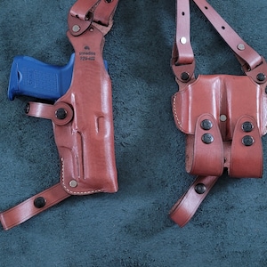 May include: Two brown leather holsters with shoulder straps. One holster is designed for a handgun and has the text "armadillo 92W-402" printed on it. The other holster is designed for two handguns.