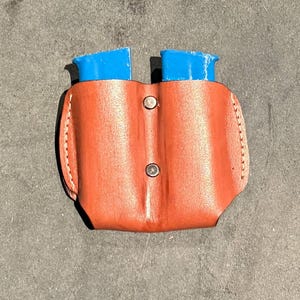 Może przedstawiać: Brązowa skórzana podwójna kabura na magazynki z dwoma niebieskimi uchwytami na magazynki. Kabura ma przeszycia na krawędziach i dwa srebrne zatrzaski.