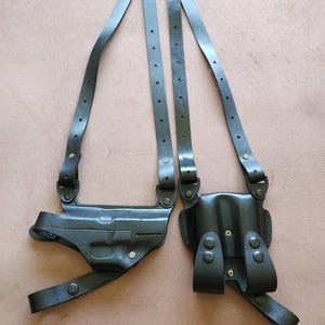May include: Black leather shoulder holster with adjustable straps and two pouches. The holster features metal rivets and a central connecting piece. The design is for carrying a firearm.