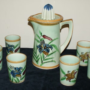 May include: A ceramic pitcher with a built-in juicer and six matching glasses. The set is light green with floral designs in blue, brown, and green. The rims of the pitcher and glasses are trimmed in brown.