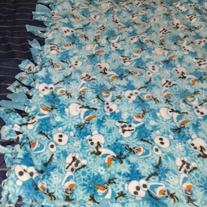 May include: A blue fleece blanket featuring a repeating pattern of Olaf the snowman from the movie Frozen, with snowflakes. The blanket has a knotted fringe border in shades of blue. A cozy and decorative item.