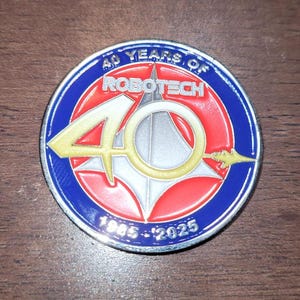 Puede incluir: Una moneda conmemorativa redonda con borde azul, centro rojo y blanco, y detalles dorados. La moneda dice "40 YEARS OF ROBOTECH" y "1985-2025". El número "40" se muestra prominentemente en dorado.