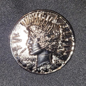May include: A silver coin with a detailed profile of a woman wearing a headdress. The coin's surface is reflective, and it features the word "LIBERTY". The coin is circular and has intricate detailing.
