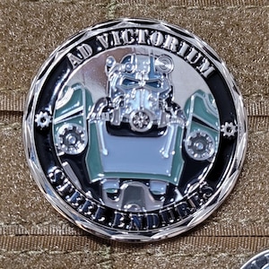 May include: A silver and black round coin featuring a green and silver robot wearing a helmet. The coin is inscribed with the text "Ad Victorium" and "Steel Endures" around the edge.
