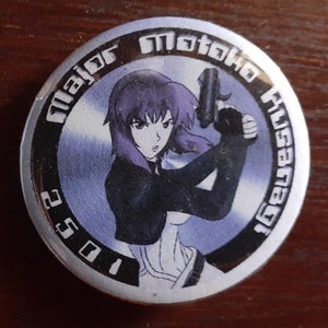 May include: A silver-colored round button with a black border and a picture of a female anime character with purple hair holding a gun. The text "Major Motoko Kusanagi" is written in a circle around the character. The number "1052" is written in the bottom left corner.