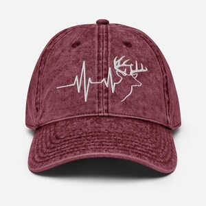 Könnte beinhalten: Burgunderfarbene Baseballkappe mit weißem Stickdesign. Das Design zeigt eine Hirschkopf-Silhouette und eine Herzschlaglinie. Die Kappe hat einen gebogenen Schirm und einen verstellbaren Riemen.