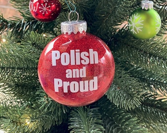 May include: A red Christmas bauble with glitter and the words "Polish and Proud" in white lettering. The bauble is hanging on a green Christmas tree with other baubles.