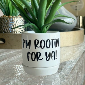 Puede incluir: Maceta de cerámica blanca con una planta suculenta verde. La maceta tiene el texto "I'm rootin' for ya!" en letras negras.