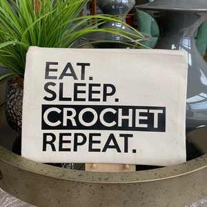 May include: A white canvas zippered pouch with black text that reads "Eat. Sleep. Crochet. Repeat."