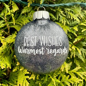 Könnte beinhalten: Runde, glitzernde Weihnachtskugel mit dem weißen Schriftzug "BEST WISHES Warmest regards". Die Kugel ist dunkelgrau mit silbernen Glitzerpartikeln und hat eine silberne Metallkappe. Dekoration für die Feiertage.