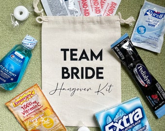May include: A white canvas bag with the text "TEAM BRIDE Hangover Kit" printed on it. The bag contains various items, including a bottle of mouthwash, a packet of electrolyte powder, a packet of vitamin C, a pack of gum, a pack of band-aids, and a hand sanitizer wipe.