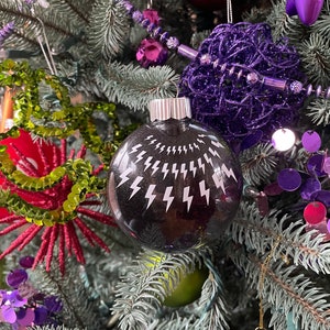 Könnte beinhalten: Ein schwarzer Weihnachtsbaumschmuck mit silbernem Glitzerfinish und weißen Blitzen. Der Schmuck hängt an einem Weihnachtsbaum mit violetten und grünen Dekorationen.