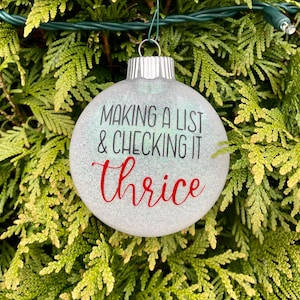 Könnte beinhalten: Eine runde, glitzernde Weihnachtskugel mit dem Text "MAKING A LIST & CHECKING IT Thrice" in Schwarz und Rot. Die Kugel hängt an einem grünen Draht vor einem Hintergrund aus grünem Laub.