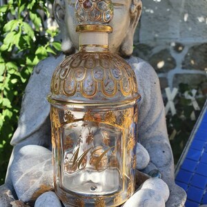 Op de afbeelding: Een doorzichtige glazen fles met een gouden dop en ingewikkelde gouden details. De fles is versierd met een patroon van bijen en de initialen "MC" zijn gegraveerd op de voorkant.