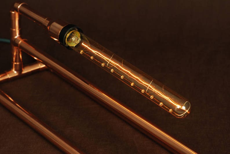 May include: A copper pipe lamp with a clear glass bulb that has a filament that looks like lightning. The bulb is lit and the light is casting a warm glow.