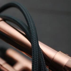 May include: Close-up of a copper pipe with black braided cable wrapped around it. The cable is woven tightly and has a smooth texture.