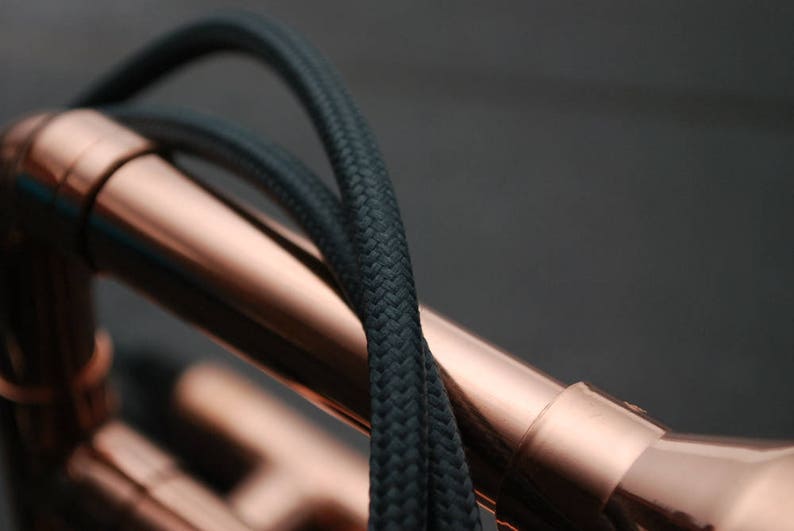 May include: Close-up of a copper pipe with black braided cable wrapped around it. The cable is woven tightly and has a smooth texture.
