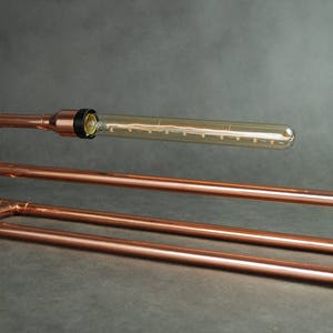 May include: A copper pipe table lamp with a clear glass bulb. The lamp has a simple, industrial design.