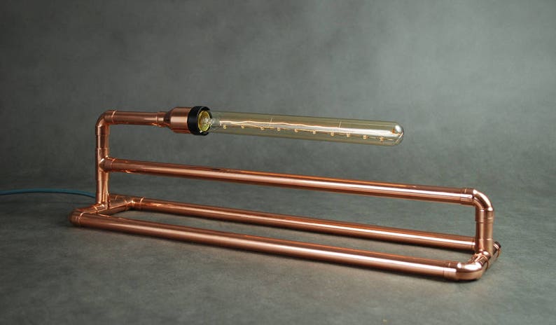May include: A copper pipe table lamp with a clear glass bulb. The lamp has a simple, industrial design.
