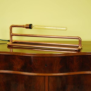 May include: A copper pipe table lamp with a clear glass bulb. The lamp is sitting on a dark wood table.