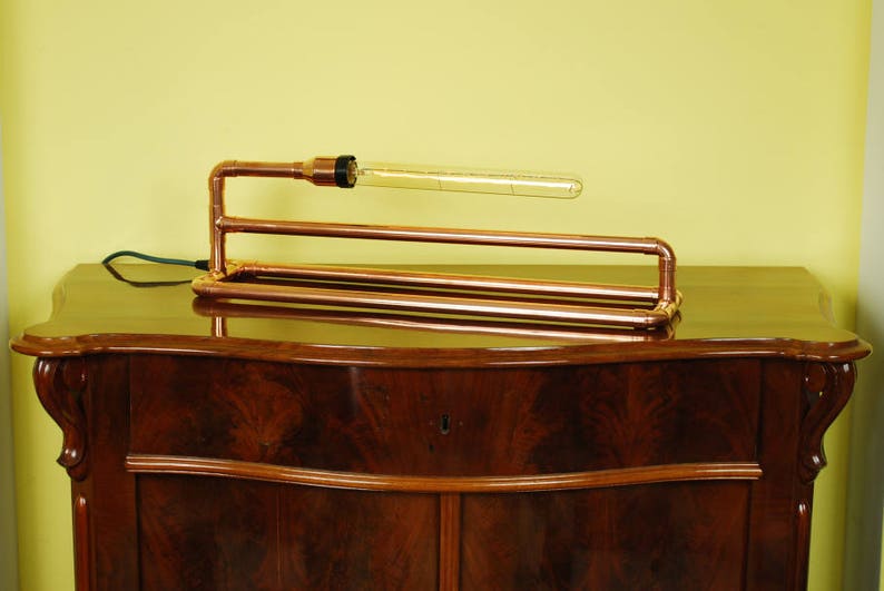 May include: A copper pipe table lamp with a clear glass bulb. The lamp is sitting on a dark wood table.