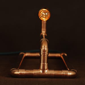 May include: A copper pipe lamp with a single light bulb. The lamp is made of copper pipes and fittings, and has a simple, industrial design.