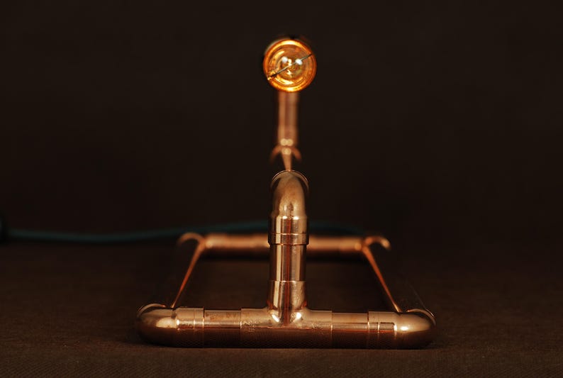May include: A copper pipe lamp with a single light bulb. The lamp is made of copper pipes and fittings, and has a simple, industrial design.