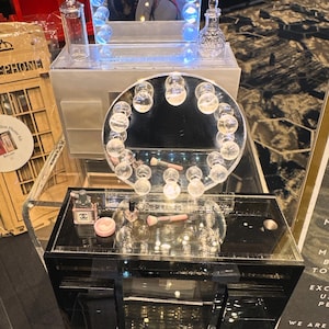 May include: A black vanity unit with a mirrored surface and a circular mirror with light bulbs. The unit has a drawer and is decorated with perfume bottles, makeup brushes, and a small blush container.