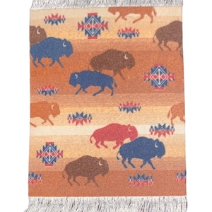 Op de afbeelding: Een geweven onderzetter met een franje rand in de kleuren bruin, beige, rood en blauw. De onderzetter heeft een herhalend patroon van gestileerde buffel silhouetten en geometrische ontwerpen.