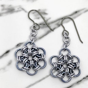 Könnte beinhalten: Ein Paar silberne und graue Ohrringe in Blumenform mit einer zarten Kette und einem Hakenverschluss.