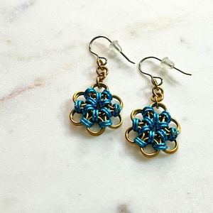Könnte beinhalten: Ein Paar handgefertigte Ohrringe mit Blumenmuster. Die Ohrringe haben einen goldfarbenen Metallrahmen mit blau gewebten Akzenten. Die Ohrhaken sind goldfarben mit transparenten Kunststoffstoppern. Die Ohrringe werden auf einer weißen Marmoroberfläche präsentiert.