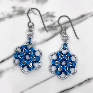 Könnte beinhalten: Ein Paar silberne Ohrringe mit blauen, blütenförmigen Anhängern. Die Ohrringe haben ein schlichtes Design und sind aus Metall.