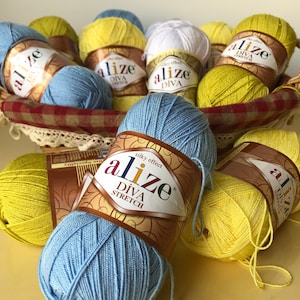 Puede incluir: Una cesta llena de ovillos de lana de varios colores, incluyendo amarillo, azul y blanco. La lana está etiquetada como "Alize Diva Stretch" con el texto "efecto sedoso" en la etiqueta.