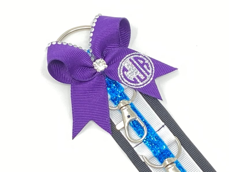 May include: A purple ribbon bow with a silver glitter monogram and rhinestones attached to a key chain with two silver swivel clips and a blue glitter ribbon.