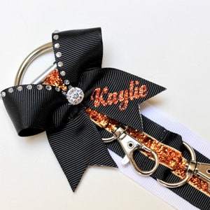 May include: Black and white ribbon with a copper glitter stripe and a black bow with rhinestones. The ribbon has two silver clips and the bow has a large rhinestone in the center. The ribbon has the name "Kaylie" written in copper glitter.