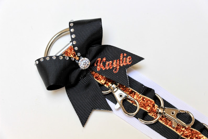 May include: Black and white ribbon with a copper glitter stripe and a black bow with rhinestones. The ribbon has two silver clips and the bow has a large rhinestone in the center. The ribbon has the name "Kaylie" written in copper glitter.